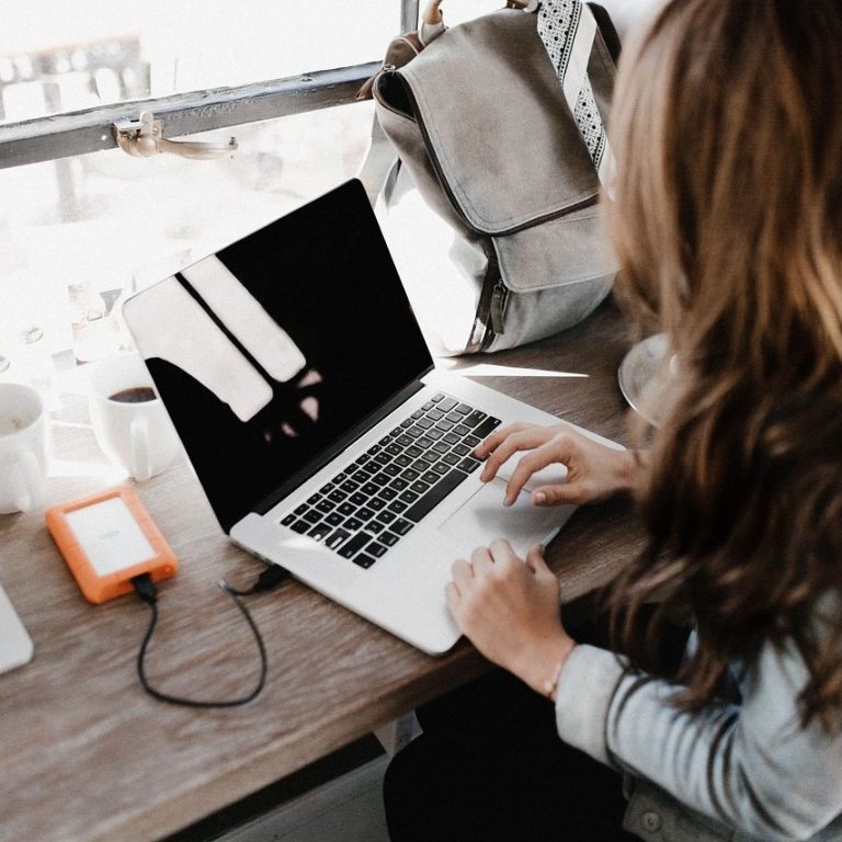 Frau tippt auf einem Laptop an einem Tisch mit einer Tasche und einer Tasse.