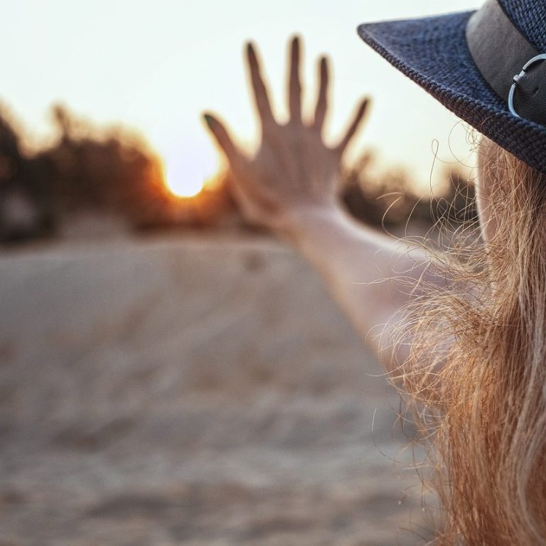 Eine Frau mit Hut winkt mit der Hand zum Sonnenuntergang.