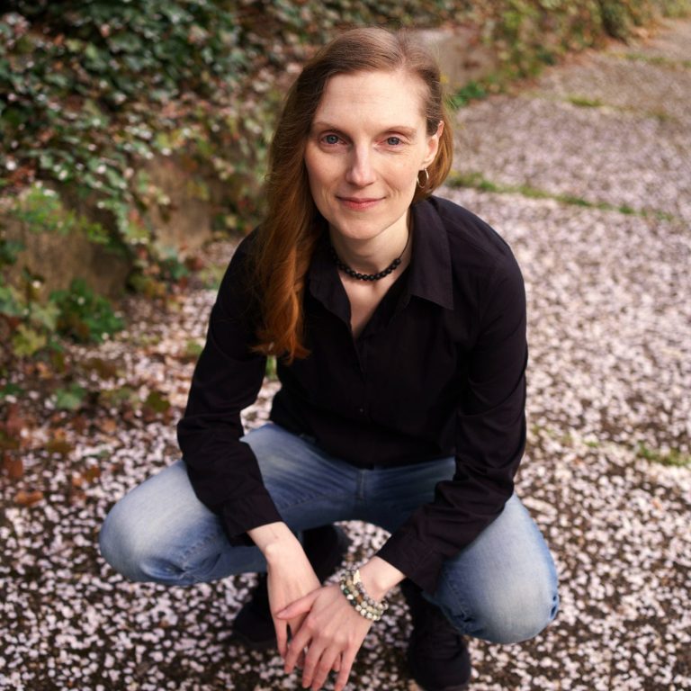 UmWegGefährtin Frau mit rotem Haar in schwarzer Lederjacke sitzt auf einem Stein in der Natur.