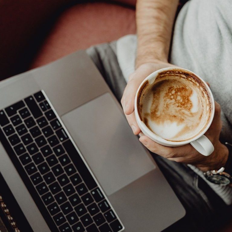 Ein Mann hält eine Tasse Kaffee über einem Laptop in beiden Händen.